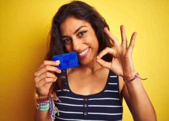 A woman with long brown hair smiles at the camera while holding a blue credit card, one of the best credit cards to build credit. She makes an "OK" gesture with her other hand. She is wearing a black and white striped tank top and has colorful bracelets on her wrist. The background is bright yellow. | FintechZoom