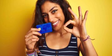 A woman with long brown hair smiles at the camera while holding a blue credit card, one of the best credit cards to build credit. She makes an "OK" gesture with her other hand. She is wearing a black and white striped tank top and has colorful bracelets on her wrist. The background is bright yellow. | FintechZoom