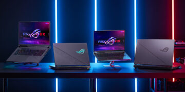 A row of four ASUS ROG laptops, touted as the best gaming laptops, is displayed on a table against a backdrop of blue and red vertical neon lights. The laptops are open, showcasing futuristic screens with the ROG logo. The ambient light creates a high-tech, gaming atmosphere. | FintechZoom