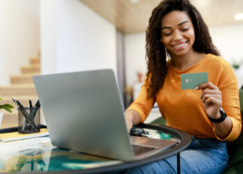 A woman with long curly hair, wearing an orange shirt, sits at a table with a laptop, holding a green bank card in her hand. She is smiling and appears to be shopping online for the best credit cards. The table also has a notepad, pen holder, and a pair of glasses. | FintechZoom