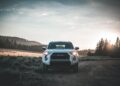 A white Toyota SUV is parked on a dirt road in a countryside setting, with a forest and hilly landscape in the background. The sun is rising or setting, casting a warm glow over the scene. The sky is partly cloudy, contributing to the serene atmosphere. VIN decoding could reveal its history. | FintechZoom