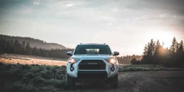 A white Toyota SUV is parked on a dirt road in a countryside setting, with a forest and hilly landscape in the background. The sun is rising or setting, casting a warm glow over the scene. The sky is partly cloudy, contributing to the serene atmosphere. VIN decoding could reveal its history. | FintechZoom