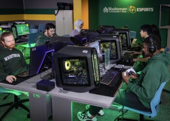 A group of people sits at computer desks in a room labeled "Washtenaw Community College ESports." They are focused on playing video games on high-end gaming computers. The brightly lit room is filled with gaming peripherals and accessories, capturing the essence of college esports competition. | FintechZoom