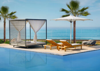 A serene poolside scene featuring a modern cabana with white drapes, two wooden lounge chairs with yellow cushions, and a white umbrella. The pool reflects the clear blue sky, and the background shows the ocean with a horizon, tropical plants, and palm trees—a perfect spot to use your hotel points. | FintechZoom