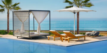 A serene poolside scene featuring a modern cabana with white drapes, two wooden lounge chairs with yellow cushions, and a white umbrella. The pool reflects the clear blue sky, and the background shows the ocean with a horizon, tropical plants, and palm trees—a perfect spot to use your hotel points. | FintechZoom