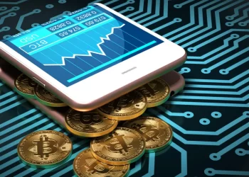 A smartphone displaying a financial chart is resting on a pile of gold bitcoins. The background features a network of intricate, blue circuit board lines, emphasizing the connection between digital currency and crypto wallets in modern technology. | FintechZoom