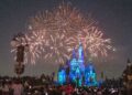 Fireworks light up the night sky above an illuminated castle, with vibrant explosions in various colors. A large crowd captures the spectacle on their devices, resembling a bustling FTSE 100 trading floor. Decorative lights and lampposts frame the foreground, evoking a festive and magical atmosphere. | FintechZoom