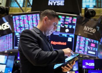 A trader, dressed in a dark suit, uses a tablet on the trading floor of the New York Stock Exchange. Multiple large screens displaying stock market information, including "Dow Jones Today," surround him, with the NYSE logo prominently visible at the top. | FintechZoom