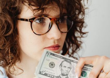 A person with curly hair and large glasses is holding a five-dollar bill close to their mouth. They appear to be in deep thought. The background is plain and out of focus, emphasizing the person's contemplative expression and the bill. | FintechZoom