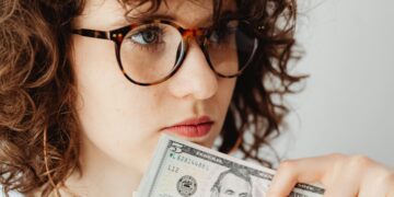 A person with curly hair and large glasses is holding a five-dollar bill close to their mouth. They appear to be in deep thought. The background is plain and out of focus, emphasizing the person's contemplative expression and the bill. | FintechZoom