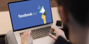 A person is typing on a laptop that prominently displays the Facebook Ads logo, which features the text "facebook ADS" next to an illustration of a hand holding a megaphone. | FintechZoom