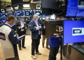 Traders on the floor of the New York Stock Exchange (NYSE) look at screens and tablets, surrounded by monitors displaying stock information and the NYSE logo. Some traders stand while others are seated, all intensely focused on their devices, analyzing the latest Markets Outlook. | FintechZoom