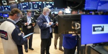 Traders on the floor of the New York Stock Exchange (NYSE) look at screens and tablets, surrounded by monitors displaying stock information and the NYSE logo. Some traders stand while others are seated, all intensely focused on their devices, analyzing the latest Markets Outlook. | FintechZoom