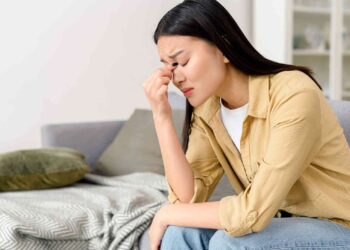 A woman in a yellow shirt and jeans sits on a couch with her eyes closed, pinching the bridge of her nose in apparent frustration or stress, perhaps worrying about financial stability. A gray blanket and green pillow are on the couch next to her, and a white shelf with books is in the background. | FintechZoom