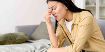 A woman in a yellow shirt and jeans sits on a couch with her eyes closed, pinching the bridge of her nose in apparent frustration or stress, perhaps worrying about financial stability. A gray blanket and green pillow are on the couch next to her, and a white shelf with books is in the background. | FintechZoom