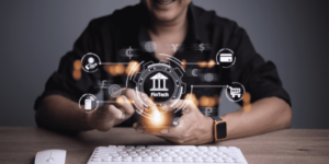 A person is sitting at a desk, holding a smartphone. Various Fintech icons, such as currency symbols, a credit card, and a shopping cart, hover above the phone, illustrating digital financial services. A keyboard is on the desk in the foreground. | FintechZoom