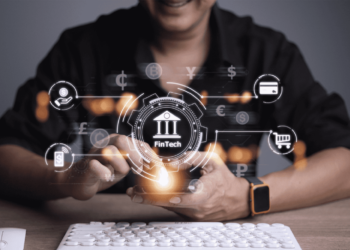 A person is sitting at a desk, holding a smartphone. Various Fintech icons, such as currency symbols, a credit card, and a shopping cart, hover above the phone, illustrating digital financial services. A keyboard is on the desk in the foreground. | FintechZoom
