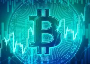 A glowing blue Bitcoin logo is prominently displayed in the center of the image against a background of rising and falling stock market charts. The image symbolizes the fluctuations in the value of Bitcoin, hinting at potential trends of Bitcoin going up among other cryptocurrencies. | FintechZoom