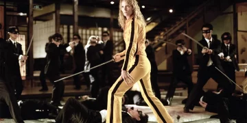 A person in a yellow tracksuit stands confidently with a sword in hand, reminiscent of one of the best movies on Netflix, surrounded by multiple individuals in black suits with blindfolds. They appear to have been defeated in combat, either lying on the ground or posed defensively, within a traditional interior setting. | FintechZoom