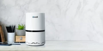 A Levoit air purifier, renowned as the best air purifier, sits on a white marble countertop. The purifier features a sleek, cylindrical design with a white body and black accents. Beside it, there is a small green potted plant and a wooden box with books and pens in the background. | FintechZoom