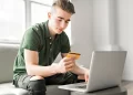 A young man in a green polo shirt is sitting on a light gray sofa using a laptop on a small table. He holds one of the best credit cards for beginners in his left hand and types on the laptop with his right hand. The background features large windows and a well-lit, clean space. | FintechZoom