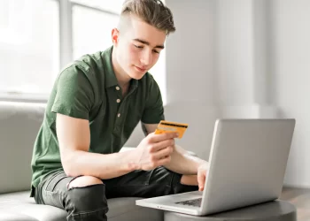 A young man in a green polo shirt is sitting on a light gray sofa using a laptop on a small table. He holds one of the best credit cards for beginners in his left hand and types on the laptop with his right hand. The background features large windows and a well-lit, clean space. | FintechZoom