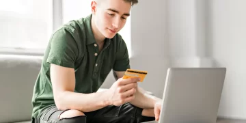 A young man in a green polo shirt is sitting on a light gray sofa using a laptop on a small table. He holds one of the best credit cards for beginners in his left hand and types on the laptop with his right hand. The background features large windows and a well-lit, clean space. | FintechZoom