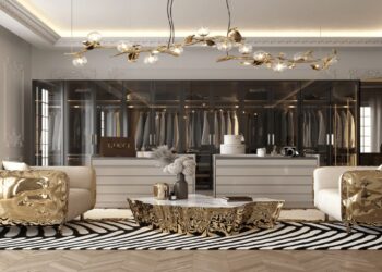 Luxurious home dressing room with a modern design. Features plush white seating with metallic gold accents, a central marble table with intricate gold detailing, and an opulent chandelier with branch-like elements and exposed bulbs. A glass wardrobe showcases hanging clothes.
 | FintechZoom