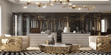 Luxurious home dressing room with a modern design. Features plush white seating with metallic gold accents, a central marble table with intricate gold detailing, and an opulent chandelier with branch-like elements and exposed bulbs. A glass wardrobe showcases hanging clothes.
 | FintechZoom