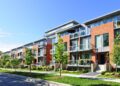 A modern multifamily property featuring a brick exterior, large windows, and balconies. The buildings are surrounded by well-maintained lawns, young trees, and walkways. The sky is clear and blue, indicating a sunny day. | FintechZoom