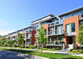 A modern multifamily property featuring a brick exterior, large windows, and balconies. The buildings are surrounded by well-maintained lawns, young trees, and walkways. The sky is clear and blue, indicating a sunny day. | FintechZoom
