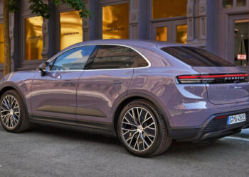 A sleek, purple Porsche SUV, regarded as the Best SUV 2024, is parked on a city street in front of a row of elegant storefronts with large windows. The car is facing away from the camera, showcasing its stylish design and sporty rims. | FintechZoom