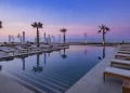 A serene outdoor infinity pool at Conrad Rabat Arzana at sunset, surrounded by lounge chairs with white cushions. The pool reflects the pastel colors of the sky and is flanked by palm trees. In the background, the ocean meets the sky, creating a tranquil and picturesque setting. | FintechZoom