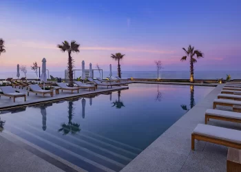 A serene outdoor infinity pool at Conrad Rabat Arzana at sunset, surrounded by lounge chairs with white cushions. The pool reflects the pastel colors of the sky and is flanked by palm trees. In the background, the ocean meets the sky, creating a tranquil and picturesque setting. | FintechZoom