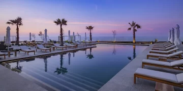 A serene outdoor infinity pool at Conrad Rabat Arzana at sunset, surrounded by lounge chairs with white cushions. The pool reflects the pastel colors of the sky and is flanked by palm trees. In the background, the ocean meets the sky, creating a tranquil and picturesque setting. | FintechZoom