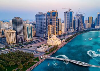 Aerial view of a coastal city featuring a line of modern high-rise buildings, showcasing prime real estate in Sharjah, a grand mosque with white domes and minarets, a pedestrian bridge extending over turquoise waters, and boats creating ripples in the harbor. Green palm trees line the waterfront. | FintechZoom