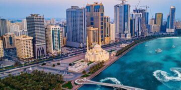 Aerial view of a coastal city featuring a line of modern high-rise buildings, showcasing prime real estate in Sharjah, a grand mosque with white domes and minarets, a pedestrian bridge extending over turquoise waters, and boats creating ripples in the harbor. Green palm trees line the waterfront. | FintechZoom