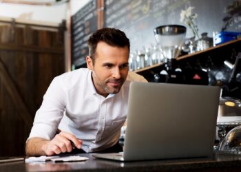 A man in a white shirt is leaning over a counter in a café, working on a laptop. Behind him, chalkboard menus and shelves with various coffee-making items are visible. He appears focused and engaged with the screen, possibly reviewing tax credits to optimize his financial planning. | FintechZoom
