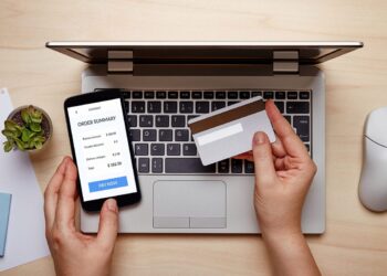 A person is holding a credit card in one hand and a smartphone displaying an order summary screen in the other. The laptop in front of them shows a keyboard and trackpad, with a small potted plant and a white computer mouse nearby on the wooden table, highlighting the need for vigilance against online payment scams. | FintechZoom