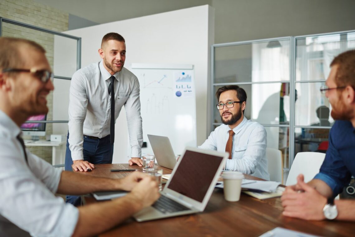 Four men in a business meeting are gathered around a wooden table with laptops and papers. One man is standing and speaking while the others are seated and listening intently. A whiteboard with charts and graphs, indicative of strategic consulting, is visible in the background, suggesting an office setting. | FintechZoom