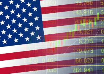 An American flag is overlaid on a background of fluctuating U.S. Stock Market numbers and financial data. The flag's stars and stripes are semi-transparent, allowing the green and yellow stock figures to be visible through the flag. | FintechZoom