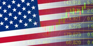 An American flag is overlaid on a background of fluctuating U.S. Stock Market numbers and financial data. The flag's stars and stripes are semi-transparent, allowing the green and yellow stock figures to be visible through the flag. | FintechZoom