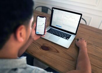 A person sits at a wooden desk using a smartphone and a laptop. Both screens show financial graphs and data, indicating they are analyzing personal finance and market trends. The person appears focused on the information displayed on the phone screen. | FintechZoom