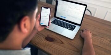 A person sits at a wooden desk using a smartphone and a laptop. Both screens show financial graphs and data, indicating they are analyzing personal finance and market trends. The person appears focused on the information displayed on the phone screen. | FintechZoom