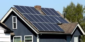 A dark blue house with brown roofing features a large array of solar panels installed on the roof, helping to reduce utility bills. There is a small chimney protruding between the panels, and a tree with green leaves is visible in the background under a clear blue sky. | FintechZoom