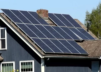 A dark blue house with brown roofing features a large array of solar panels installed on the roof, helping to reduce utility bills. There is a small chimney protruding between the panels, and a tree with green leaves is visible in the background under a clear blue sky. | FintechZoom