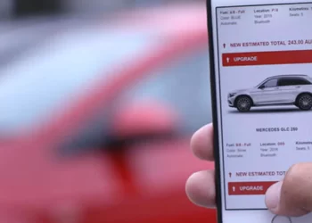 A person holding an iPhone displays a car sales app, featuring a Mercedes GLC 250. The app highlights features like full-size SUV, automatic, and a price of $243,080. In the background, a blurred red car is visible. In the latest Avis Stock update, such luxury vehicles are gaining popularity. | FintechZoom