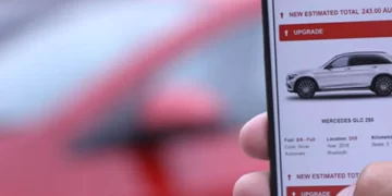 A person holding an iPhone displays a car sales app, featuring a Mercedes GLC 250. The app highlights features like full-size SUV, automatic, and a price of $243,080. In the background, a blurred red car is visible. In the latest Avis Stock update, such luxury vehicles are gaining popularity. | FintechZoom