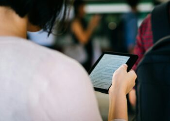 A person with short, dark hair reads from an ebook reader while standing in a crowded area. The scene is slightly blurred in the background, focusing on the device and the person's hand holding it. The person is wearing a light-colored top. | FintechZoom