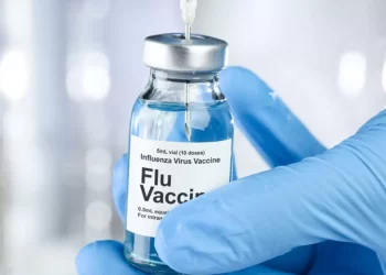 A gloved hand is holding a 5 mL vial labeled "Influenza Virus Vaccine, Flu Vaccine." A syringe needle is being inserted into the vial, drawing up the liquid vaccine. The background is blurred, highlighting the focus on the vaccine vial and syringe, reminiscent of various Moderna stock images. | FintechZoom
